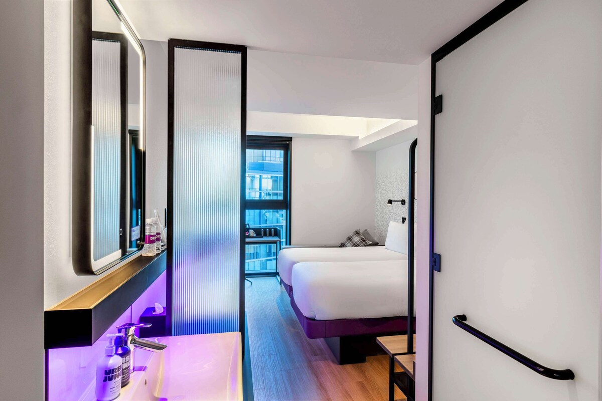A modern hotel room with a large bed, a bedside table with toiletries, a mirror, and a view of the city through a window.