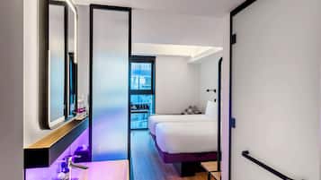 A modern hotel room with a large bed, a bedside table with toiletries, a mirror, and a view of the city through a window.