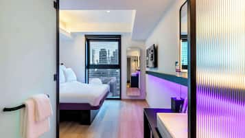 A modern hotel room with a bed, a large window, a bathroom with a mirror, and a city view.