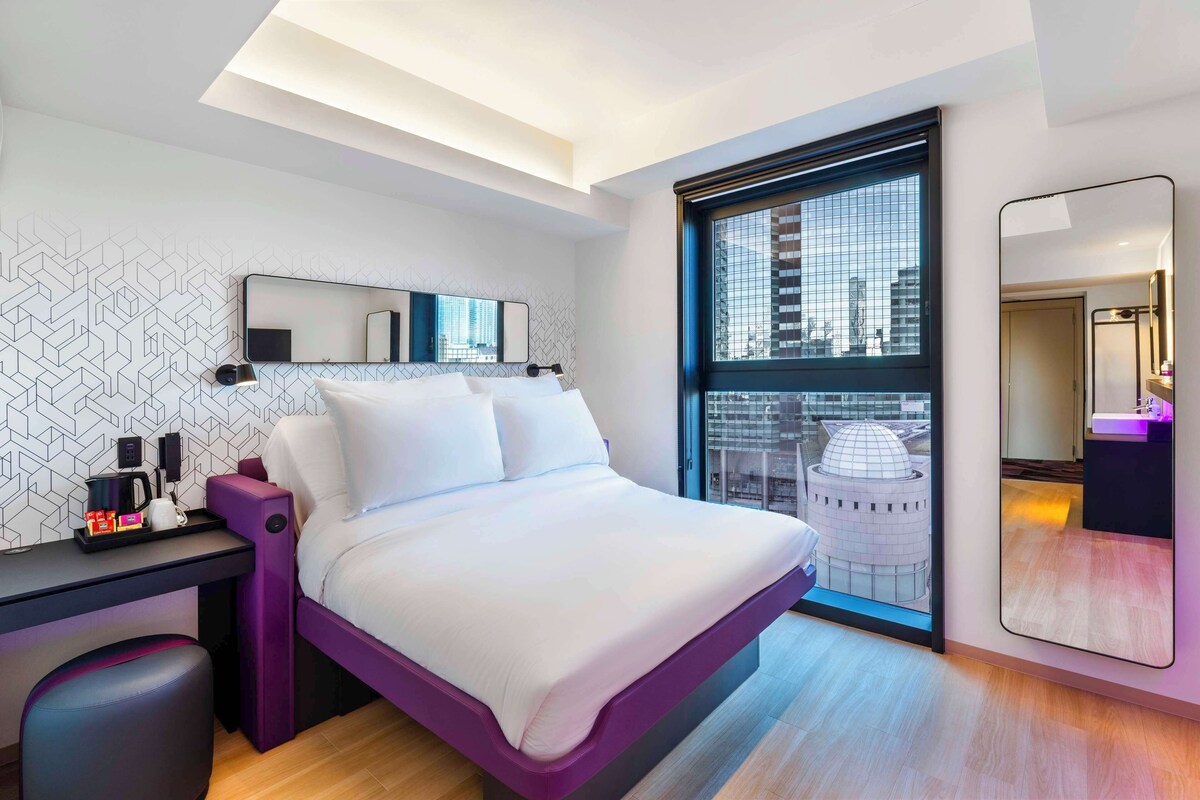 A modern hotel room with a bed, a desk, a chair, a large window with a city view, and a mirror.