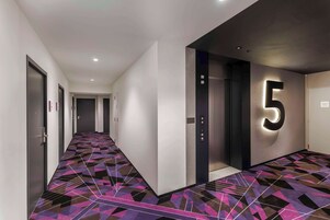 Un pasillo moderno con suelo de diseño geométrico, un elevador destacado con el número 5 y varias puertas a lo largo del corredor.