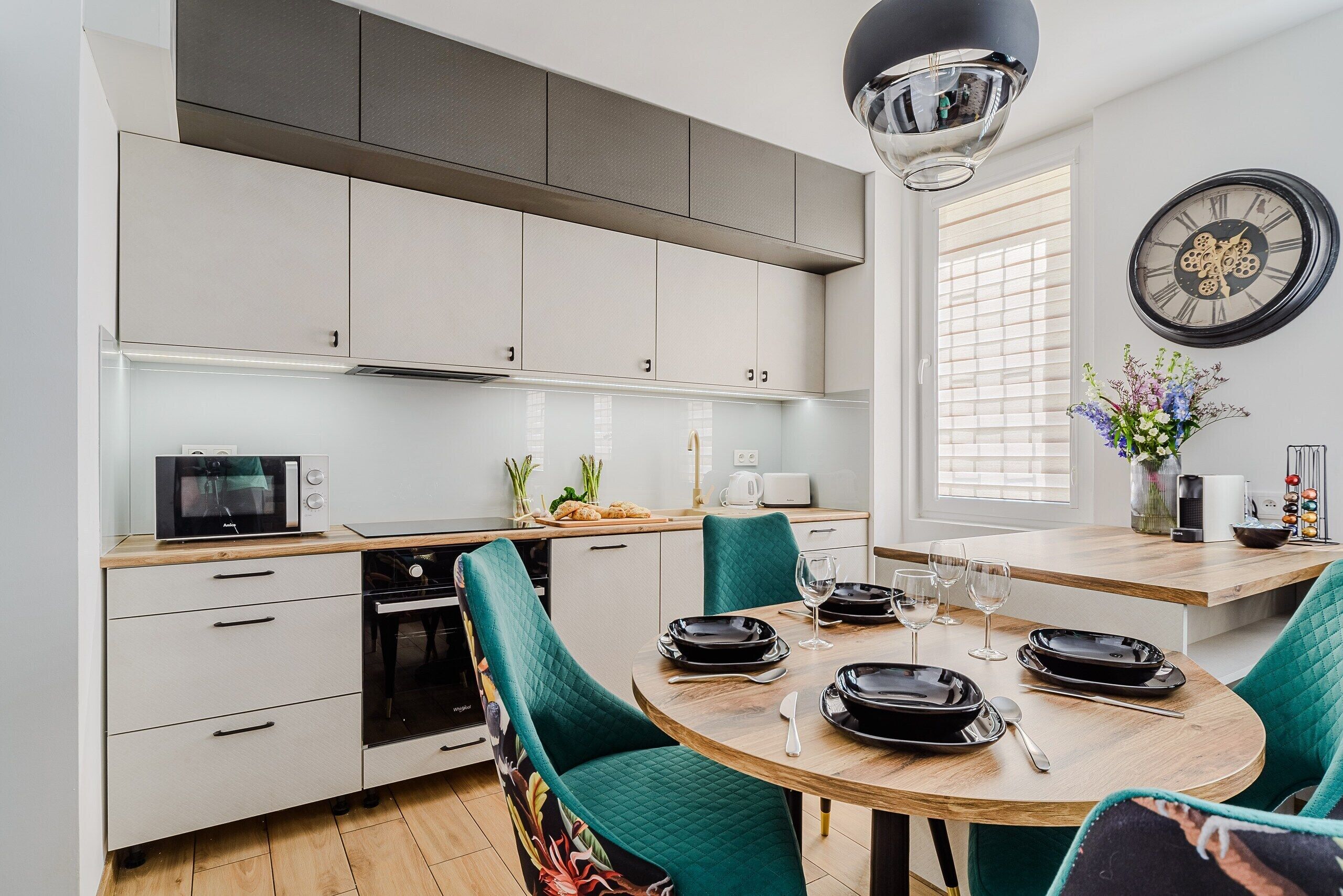Una cocina moderna con armarios blancos, microondas y un comedor con sillas color turquesa y una mesa de madera lista para dos comensales.