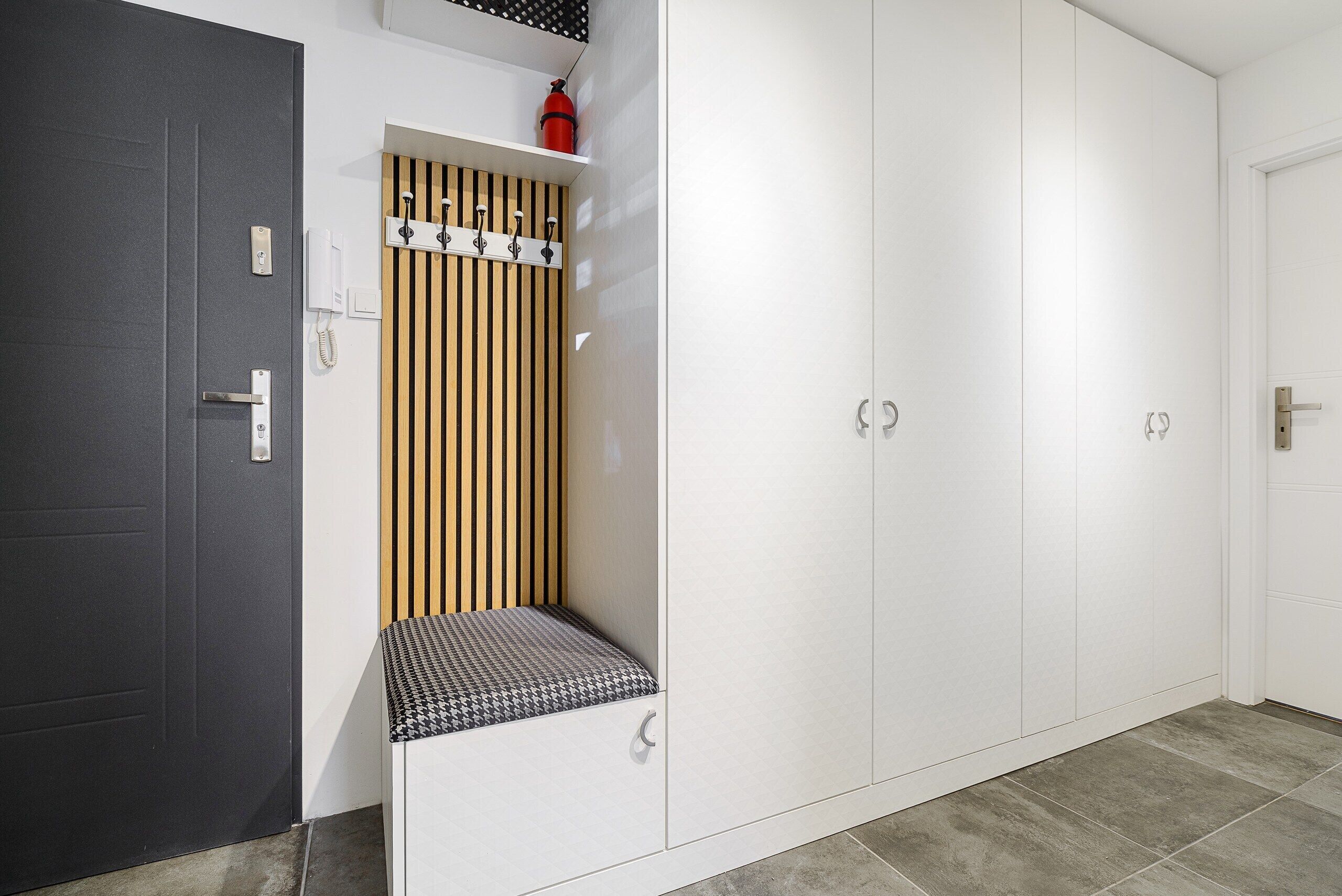Ein Flur mit einer grauen Tür, einer weißen Wand mit einem Telefon, einer Wandverkleidung aus Holzlatten, einer Bank mit gemustertem Sitz und einem weißen Kleiderschrank mit zwei Türen.