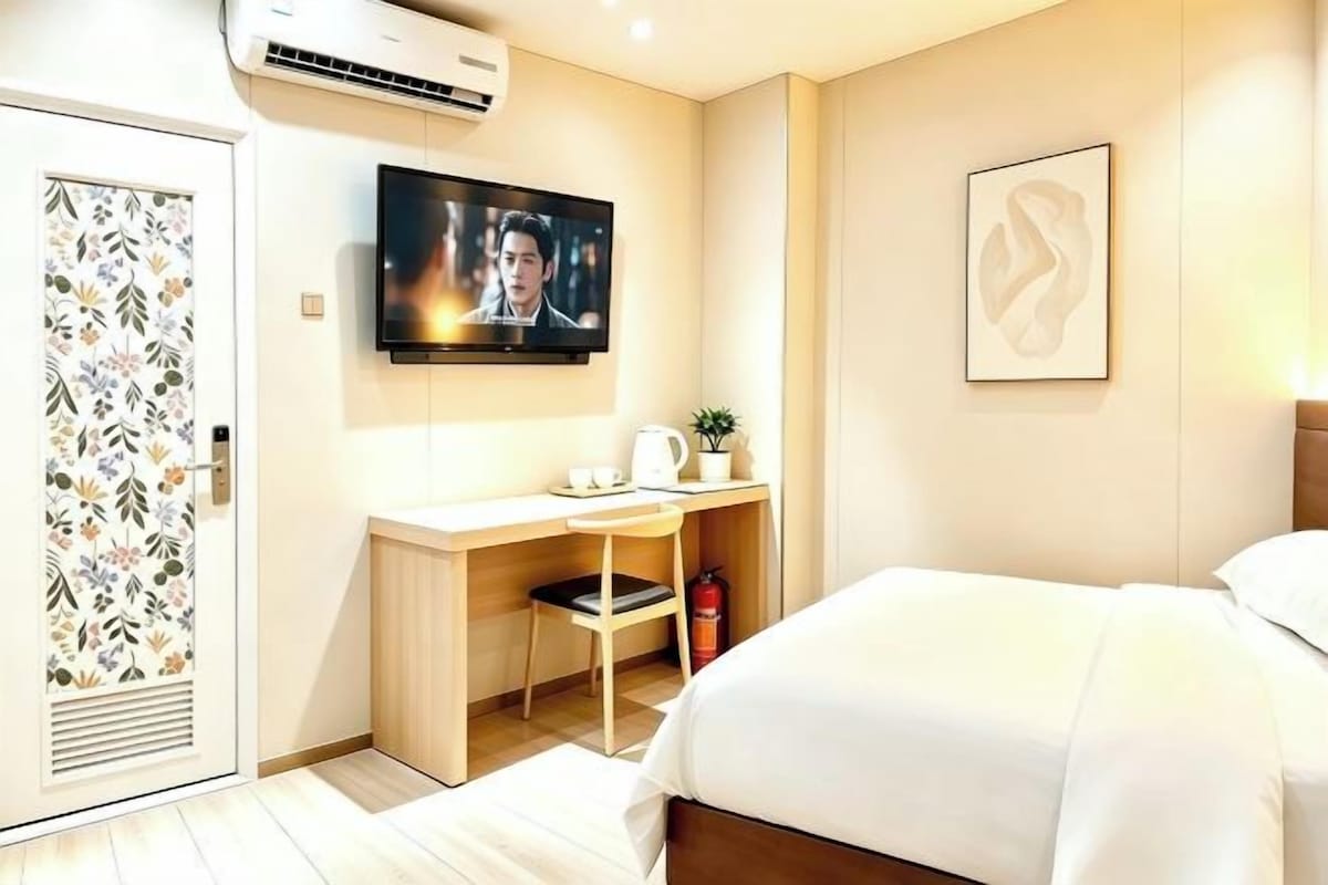 A hotel room with a bed, a desk with a chair, a television mounted on the wall, and a fire extinguisher.