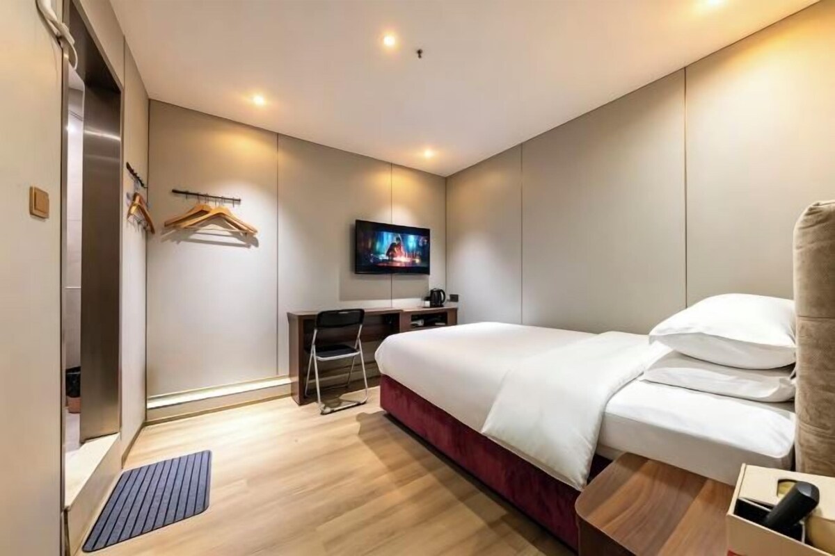 Quarto de hotel com cama, escrivaninha, cadeira, televisão e guarda-roupa.