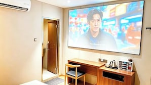 Ett hotellrum med en stor platt-TV som är monterad på väggen, ett skrivbord i trä med en stol och ett litet skåp med en vattenkokare och andra tillbehör.