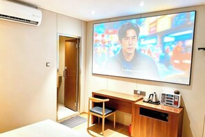 Quarto de hotel com uma grande TV de tela plana na parede, uma escrivaninha de madeira com cadeira e um pequeno armário com chaleira e outros objetos.