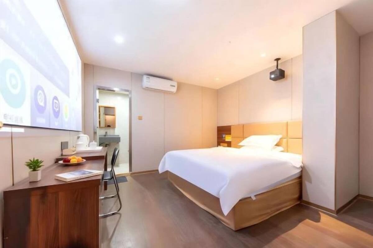 A modern hotel room with a wooden bed, a desk with a plant, and a small kitchenette.