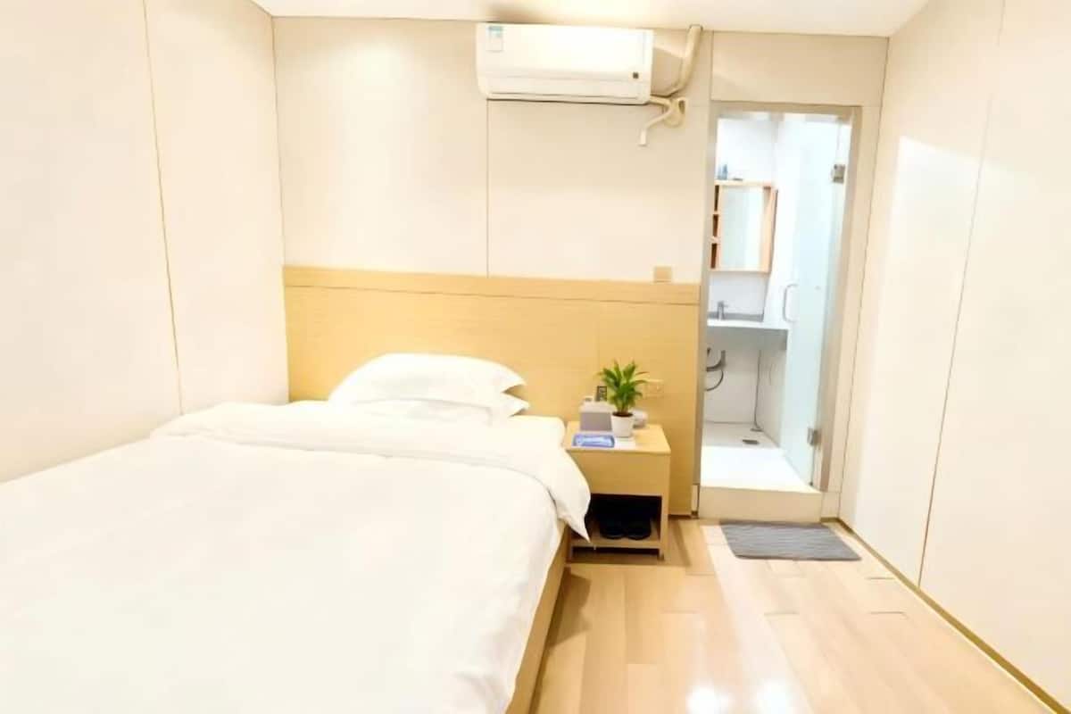A hotel room with a bed, bedside table, and a small plant. There is a door leading to a bathroom with a mirror and a shelf.
