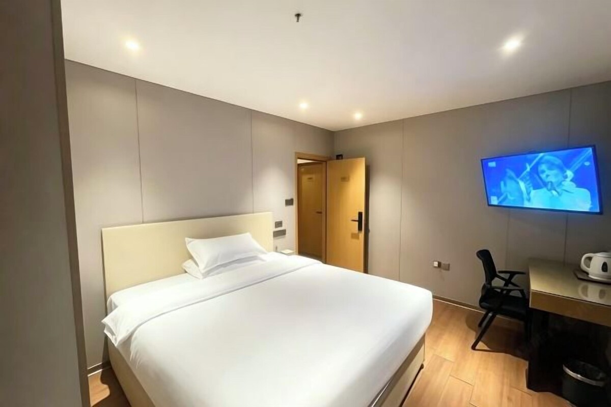 Quarto de hotel com cama, escrivaninha, cadeira e televisão na parede.