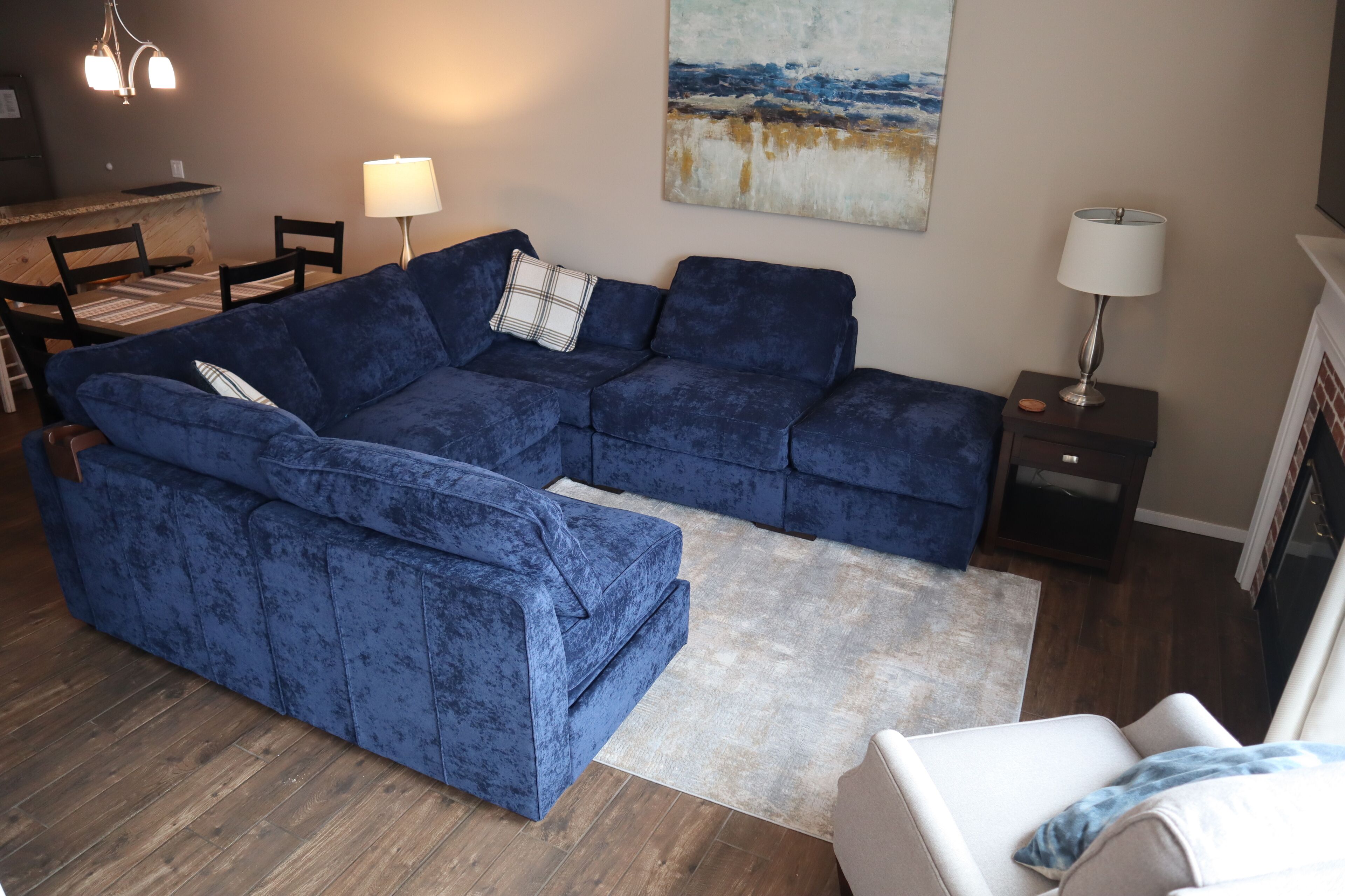 A living room with a blue sectional sofa, a dining table with chairs, a lamp, and a painting on the wall.