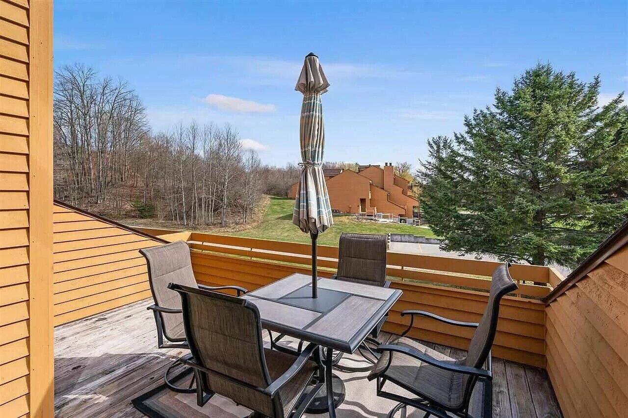 Une terrasse en bois avec une table et des chaises, un parasol et une vue sur une maison et des arbres.