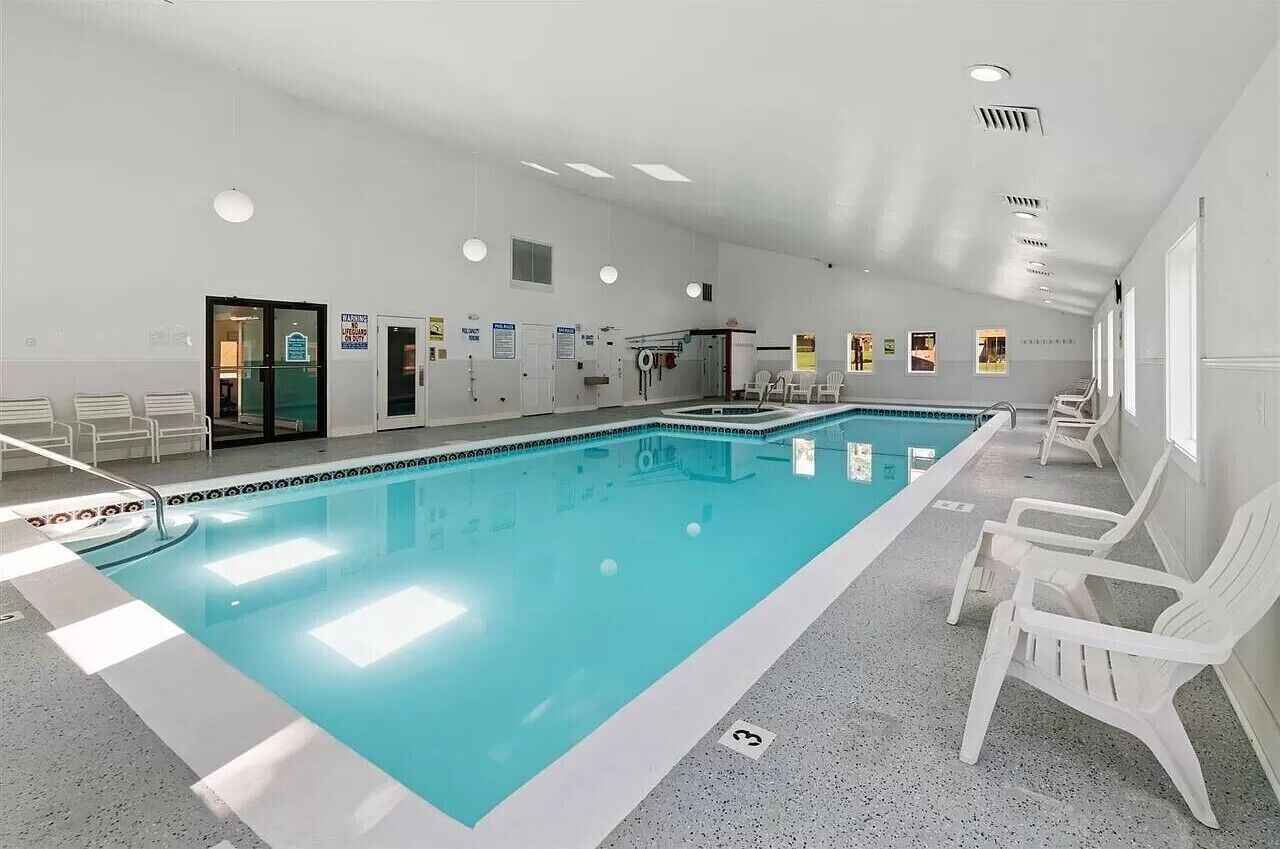 Uma grande piscina interior com superfície de água l ímpida, rodeada por cadeiras brancas e chão em mosaico.