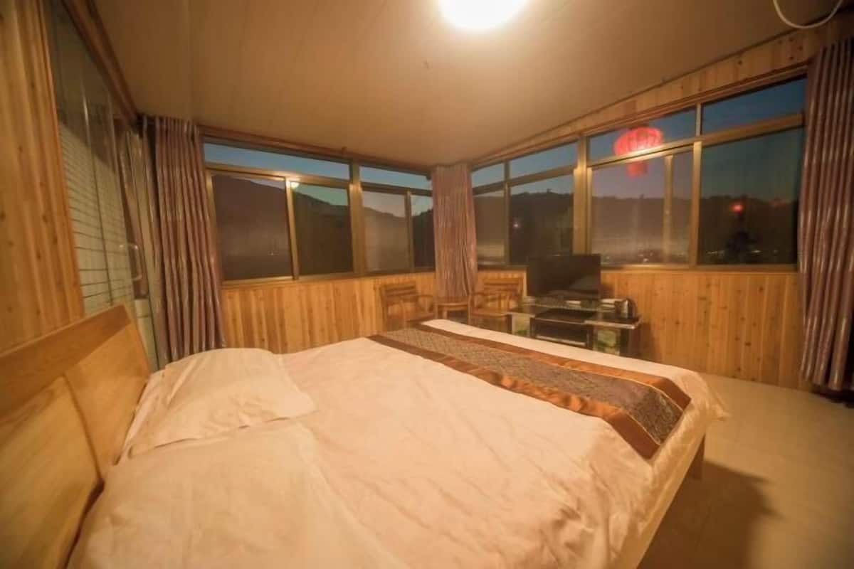 A bedroom with a bed, a desk, and a chair. There are large windows with a view of the outside.