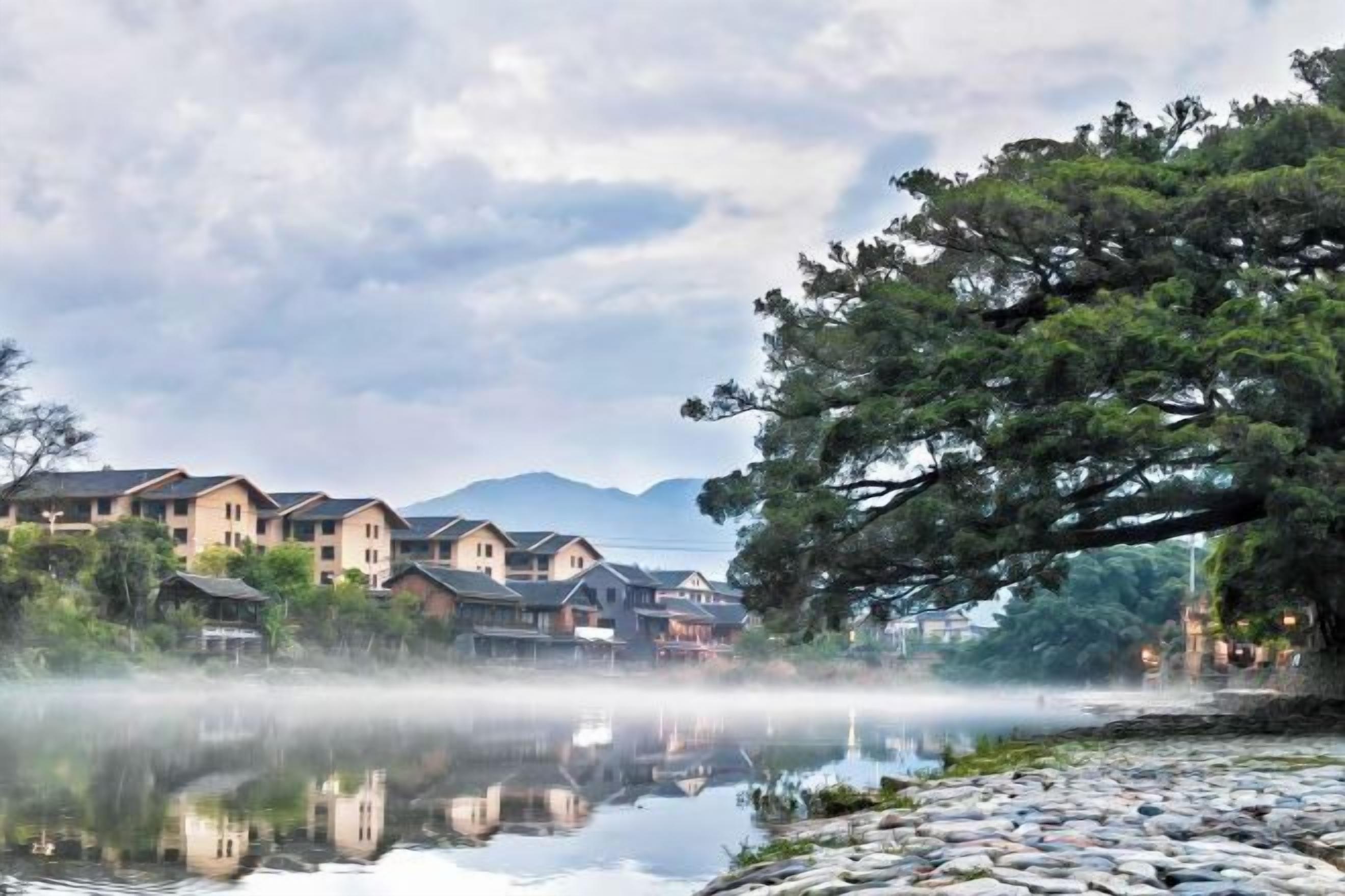 A calm lake with a reflection of buildings, a large tree, and mountains in the background.