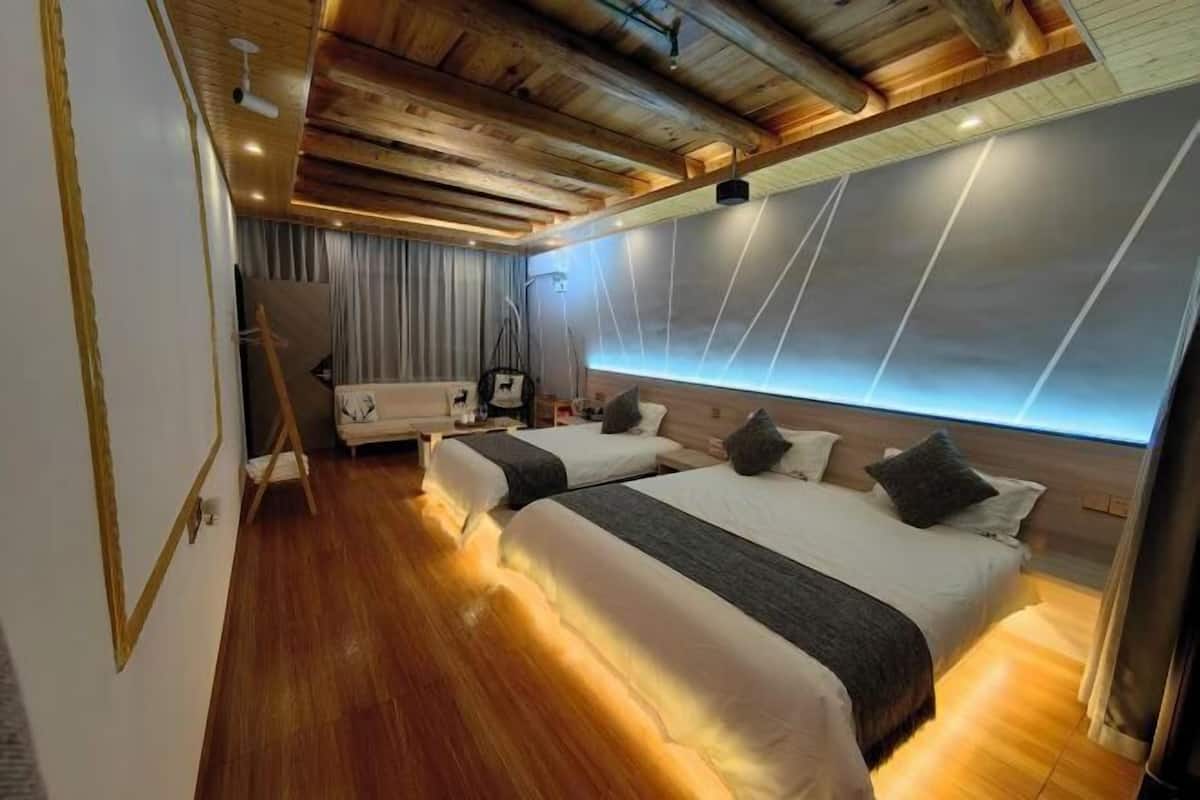 A modern hotel room with wooden flooring, two beds with grey bedding, a white sofa, and a large window with a view.