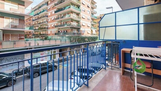 A balcony with a metal railing, a clothes drying rack, and a view of a multi-story residential building with balconies.