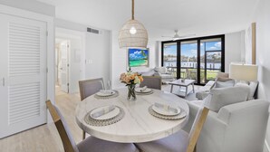 A modern dining area with a round table set for four, a vase of flowers, and a view of the outdoors through large windows. - Rare Walkout Condo | Walk to Beach & Restaurants (Marco Island)
