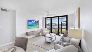 A modern living room with a large TV, comfortable seating, a coffee table, and a view of a pool and palm trees through large windows. - Rare Walkout Condo | Walk to Beach & Restaurants (Marco Island)