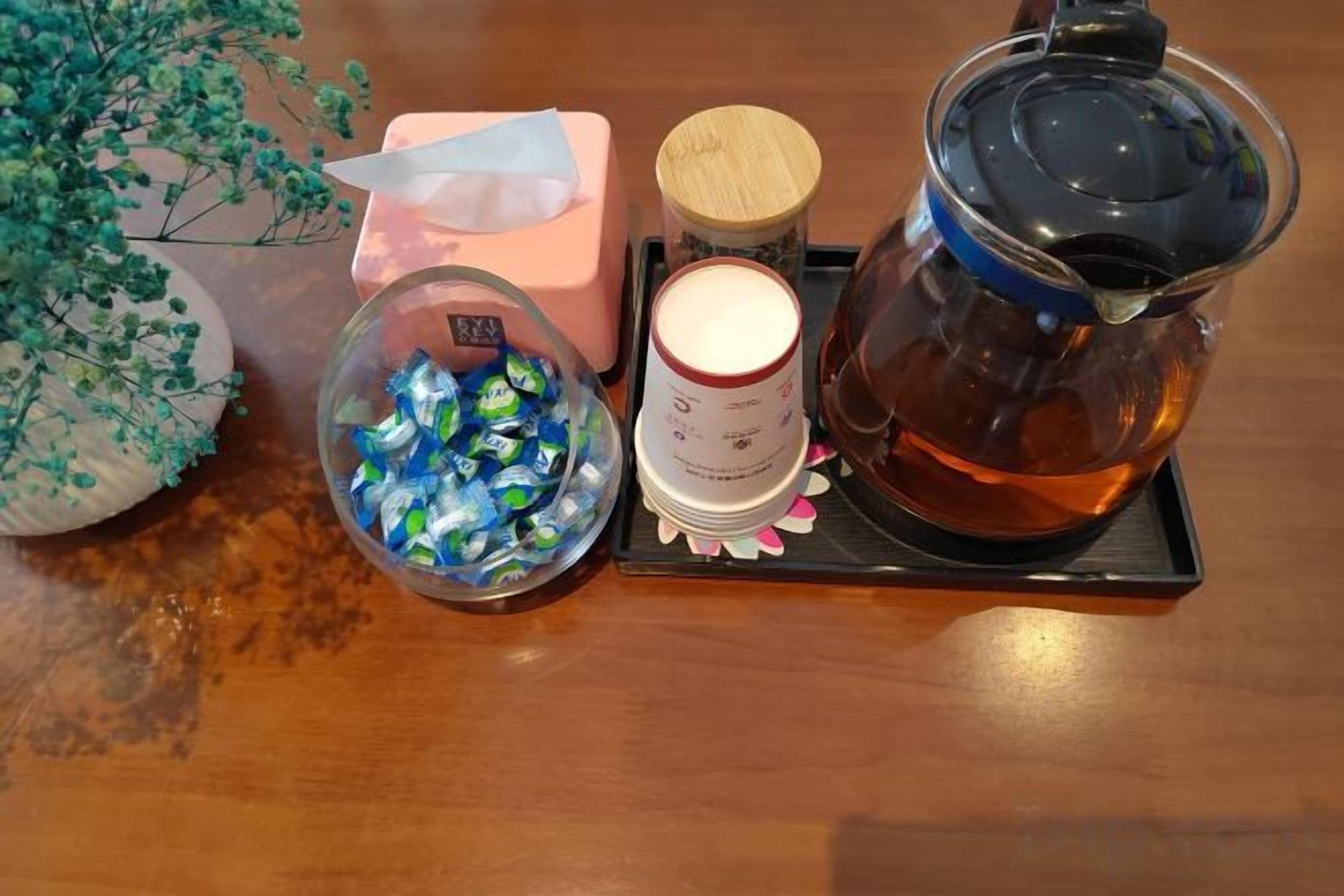 A wooden table with a tea pot, a glass jar, a tissue box, and a bowl of snacks.