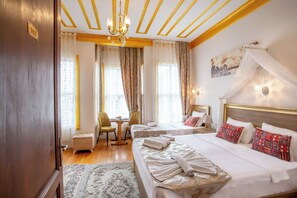 Een hotelkamer met een groot bed, twee nachtkastjes, een bureau met stoel, een klein voetenbankje en een kroonluchter.