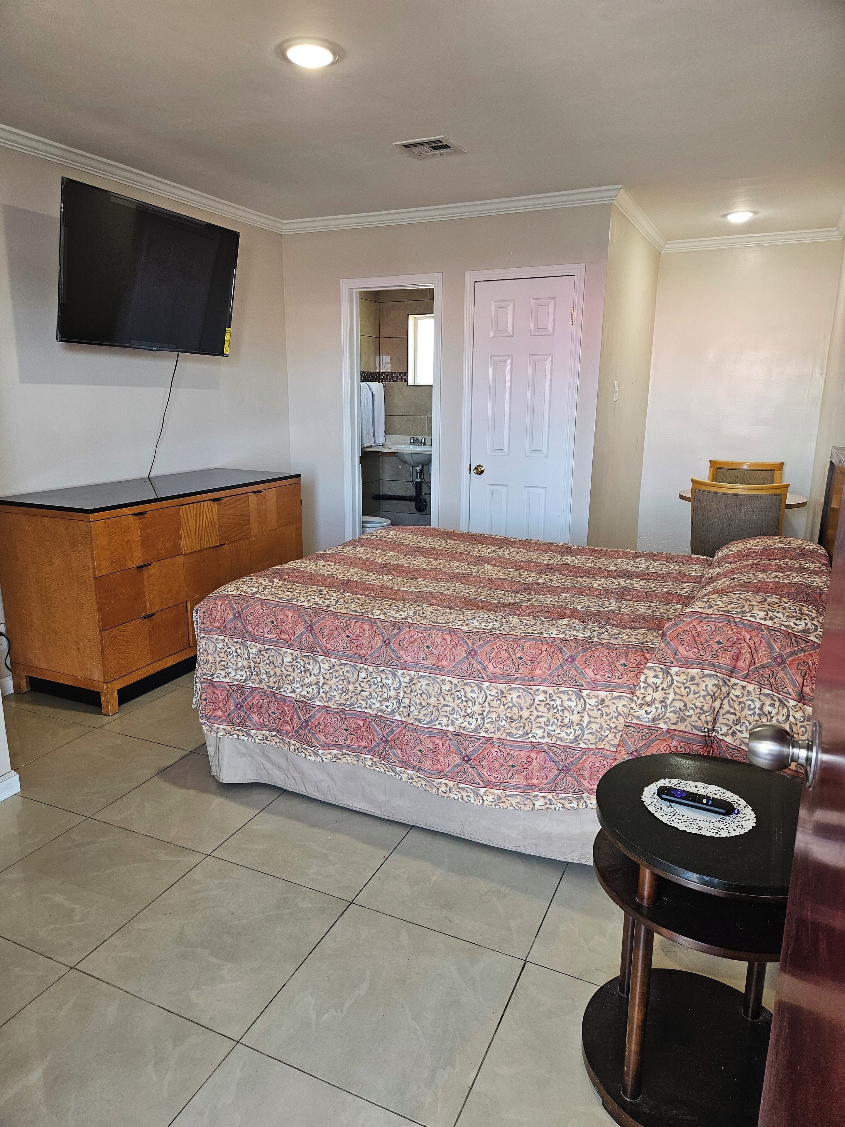 Un dormitorio con cama, cómoda, televisor montado en la pared y baño visible a través de una puerta abierta.
