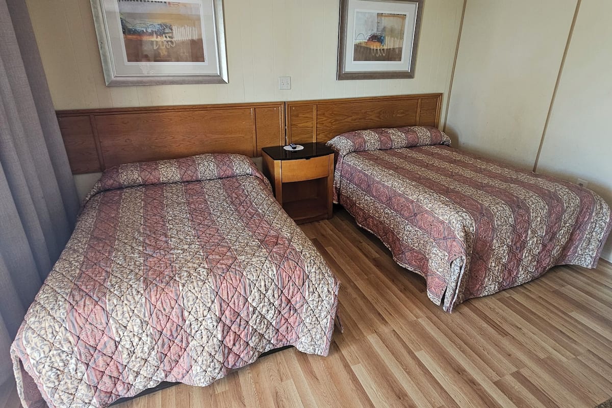 Habitación de hotel con dos camas, piso de madera, cabecero de madera, una mesita de noche y dos cuadros en la pared.