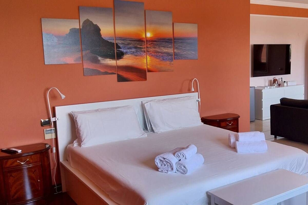 A hotel room with a large bed, a sofa, a TV, and artwork on the wall.