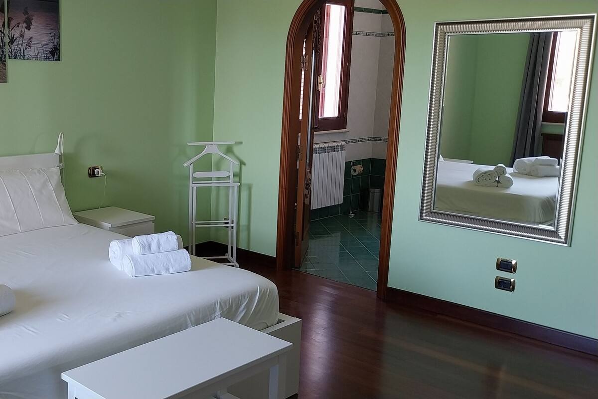 A neatly made bed with white linens and towels, a bedside table, a nightstand, a small white shelf, a mirror, and a view of a bathroom with a toilet and sink.