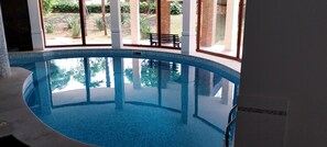 An indoor swimming pool with clear glass walls, a bench, and a view of a landscaped garden. - Villa Giulia (Syracuse)