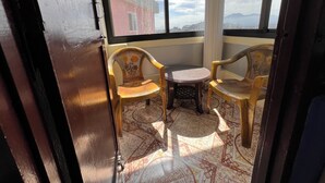 Un balcón con dos sillas de madera y una mesita pequeña, piso con diseño y vistas a edificios y montañas a través de ventanas amplias.