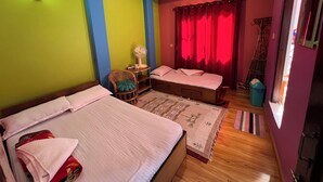Una habitación con dos camas, una silla, una mesa con un ventilador y una ventana con cortinas rojas.