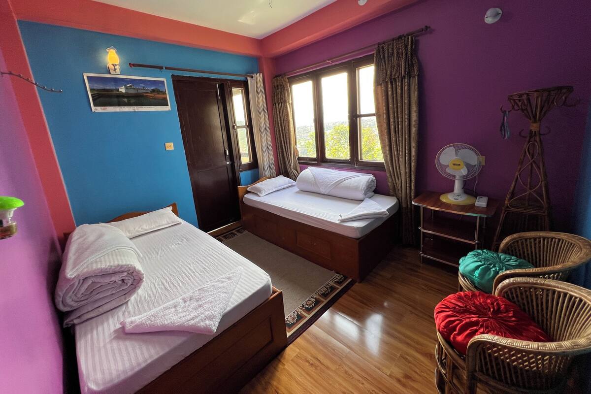 A room with two beds, a small table, a chair, a fan, and a window with curtains.