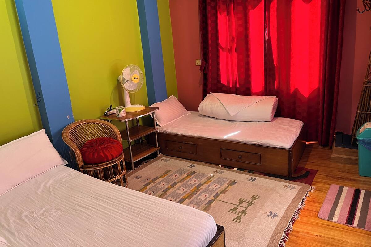 A bedroom with two beds, a chair, a fan, and a window with red curtains.