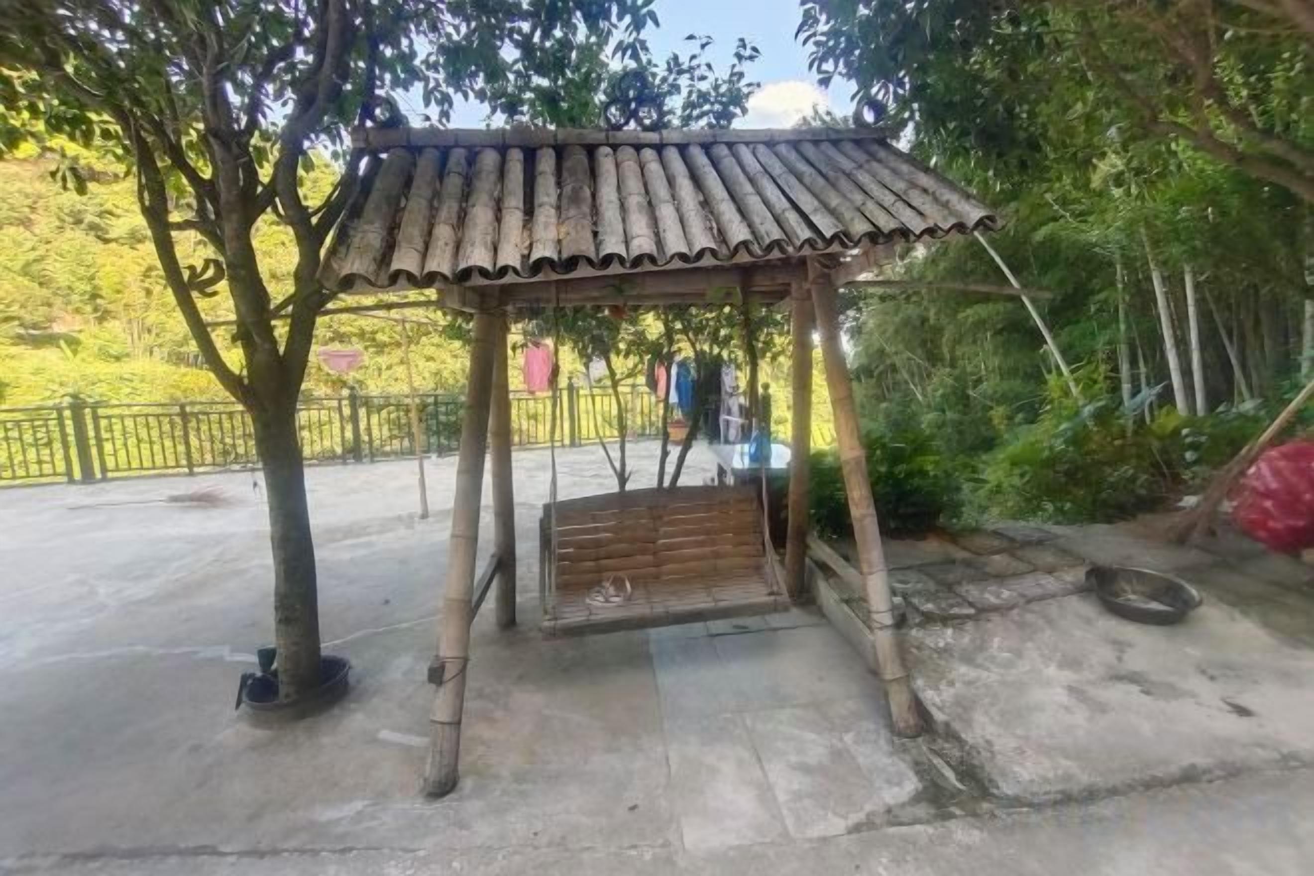A wooden gazebo with a sloped roof, surrounded by trees and a concrete ground.