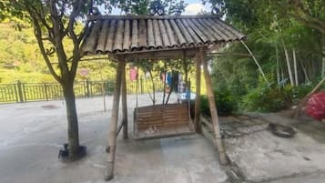 A wooden gazebo with a sloped roof, surrounded by trees and a concrete ground.