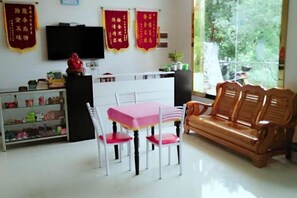 Une pièce avec un bureau blanc, une table rose et des chaises en bois. Il y a un téléviseur accroché au mur et un canapé avec des coussins.