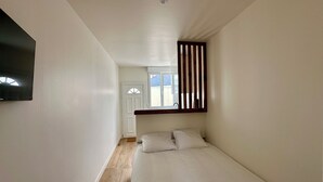 Un dormitorio con cama, ventana con persianas verticales, puerta y televisor montado.