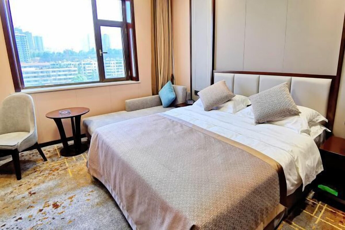 A hotel room with a large bed, a chair, a small table, and a view of the city through a large window.