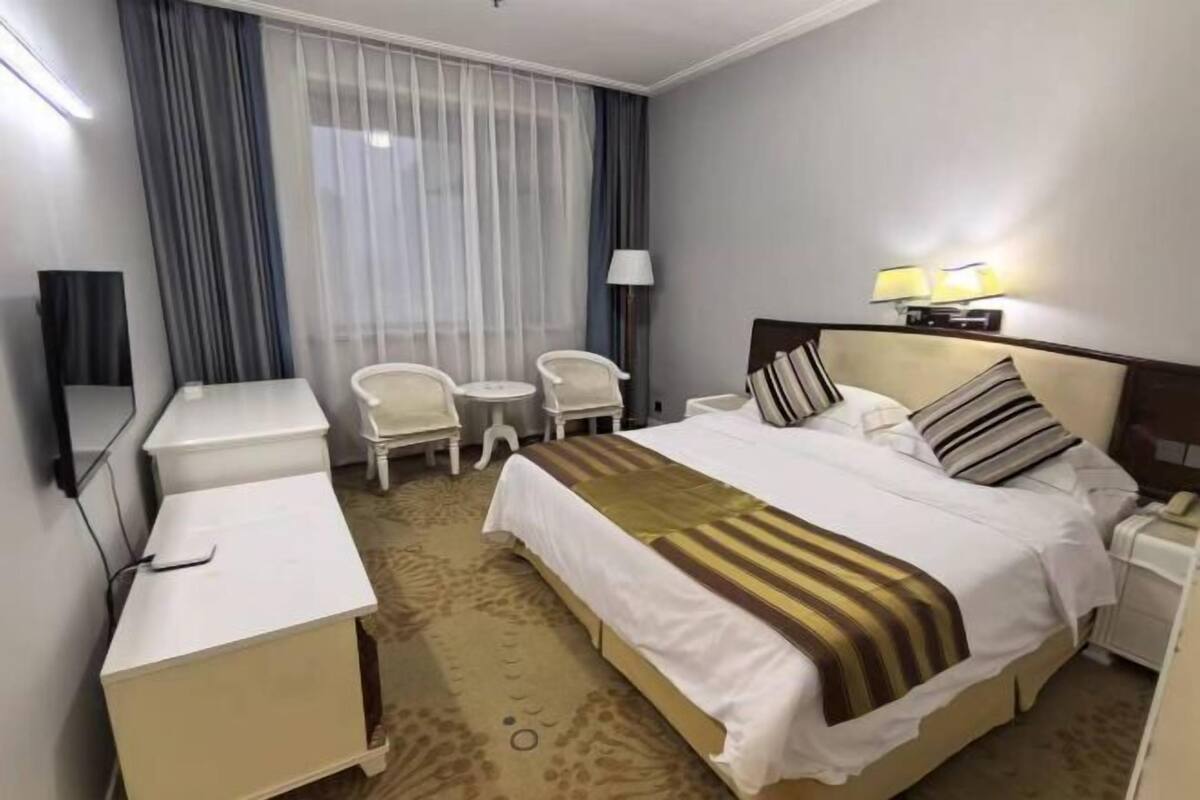 A hotel room with a bed, bedside tables, a desk, a chair, a lamp, and a window with curtains.