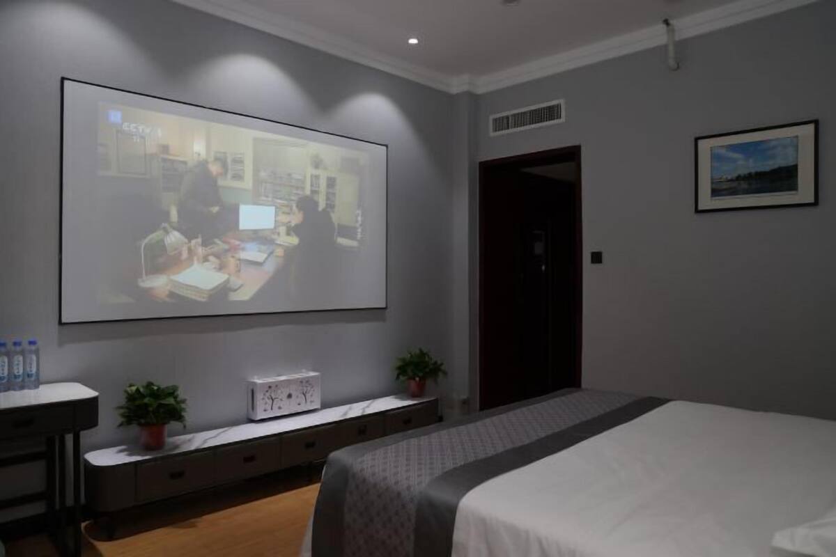 A hotel room with a bed, a desk with a computer, a projector screen, and a small plant.