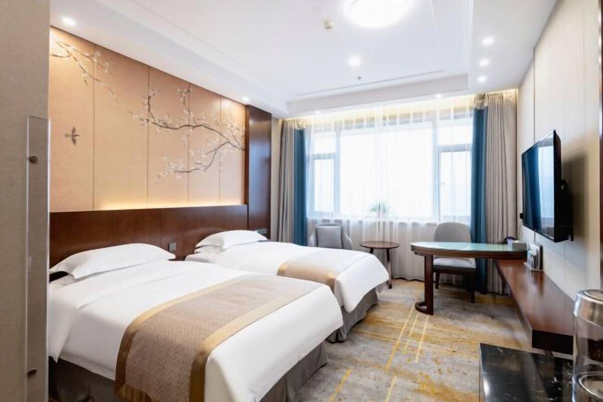 A hotel room with two beds, a desk, a chair, a TV, and a large window with curtains.