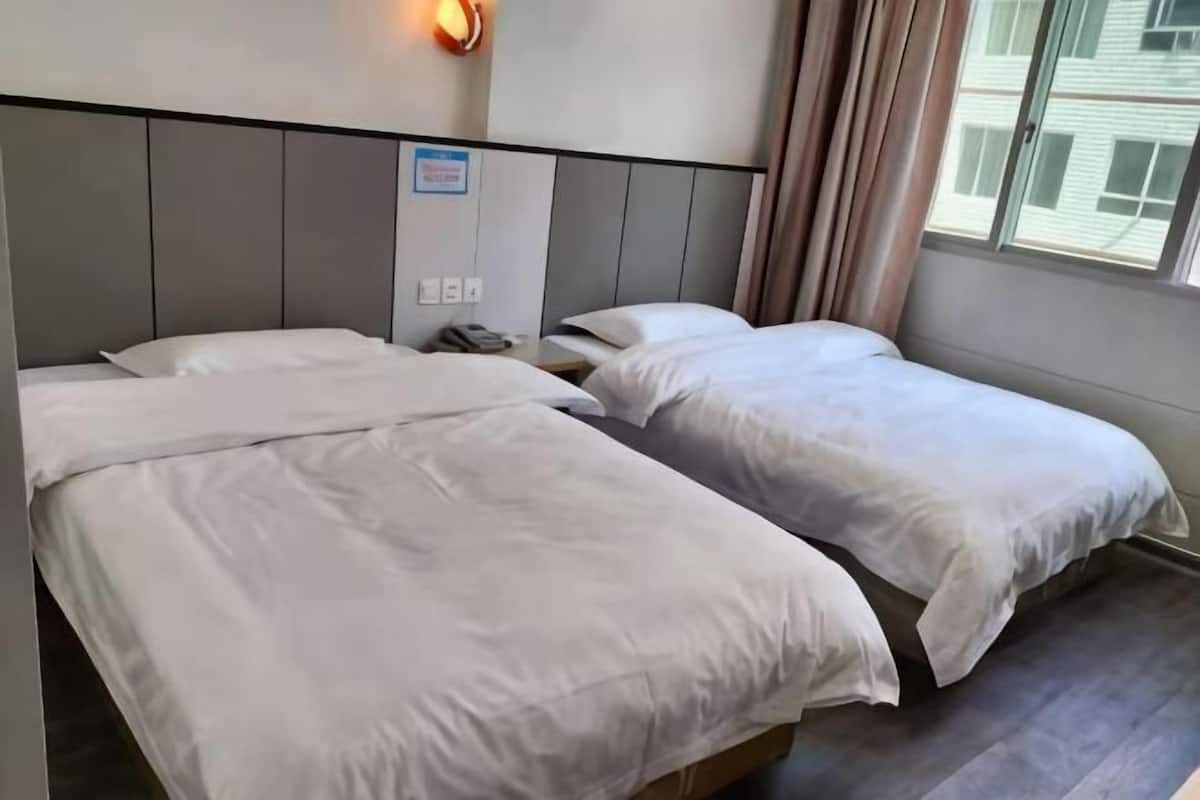 A hotel room with two beds, a nightstand, a wall-mounted light, and a window with curtains.