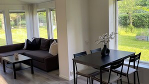 A modern living space with a sofa, coffee table, dining table, and chairs. Large windows provide views of a garden.