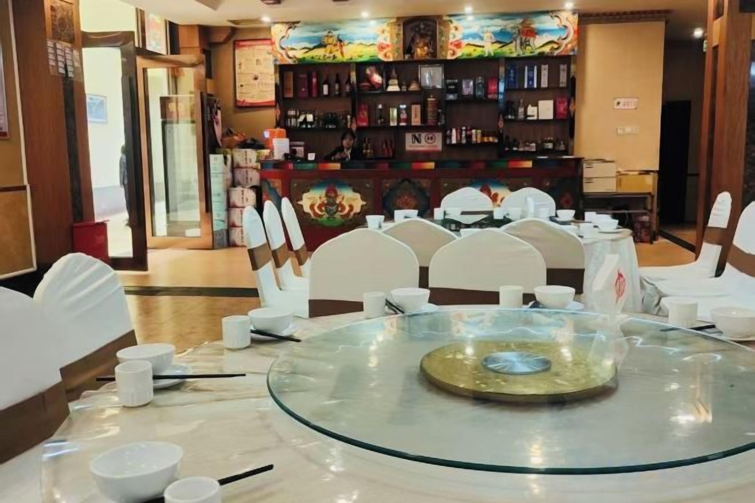 A dining area with a round glass table surrounded by white chairs, a bar area with shelves of bottles, and a colorful mural on the wall.