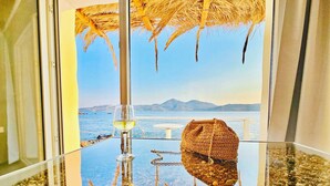 Habitación con separador de vidrio que refleja vista a la playa, una copa de vino blanco y un bolso tejido sobre una mesa.