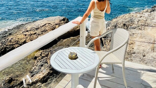 Una mujer de pie en una barandilla con vista a una zona costera, agua azul cristalina y colinas lejanas.
