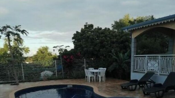 A swimming pool with a tiled deck, surrounded by a fence and trees. There are lounge chairs and a small table with chairs nearby. - 4-bedroom house for 10 people (L'Étang-Salé)