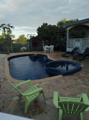Une piscine avec une terrasse carrelée, entourée d’une clôture et d’arbres. On y trouve des chaises longues et une petite table avec des chaises à proximité.