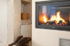 Una chimenea con fuego encendido, un estante de piedra con una cesta tejida, y un respiradero metálico debajo de la chimenea.
